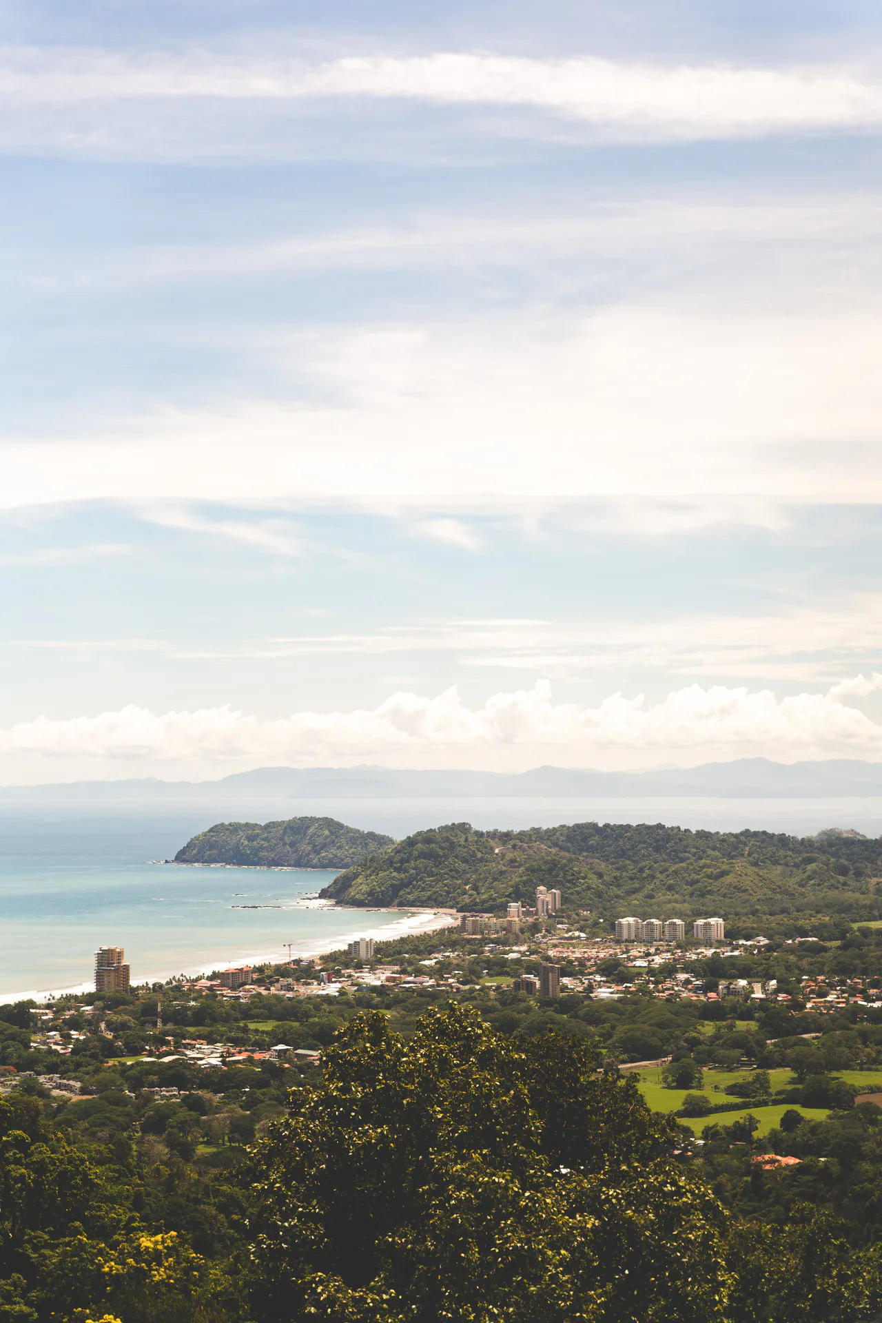 Jacó city stretches between its Pacific bay and green hills, viewed from an elevated hillside.