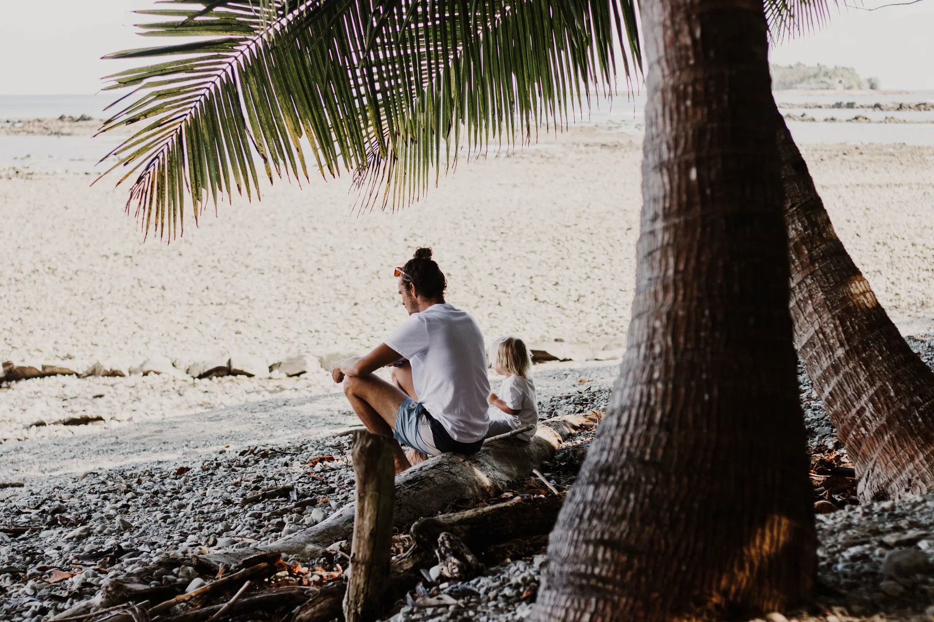 An adult and child sit together on a log in the shade of palms on a Costa Rican beach.