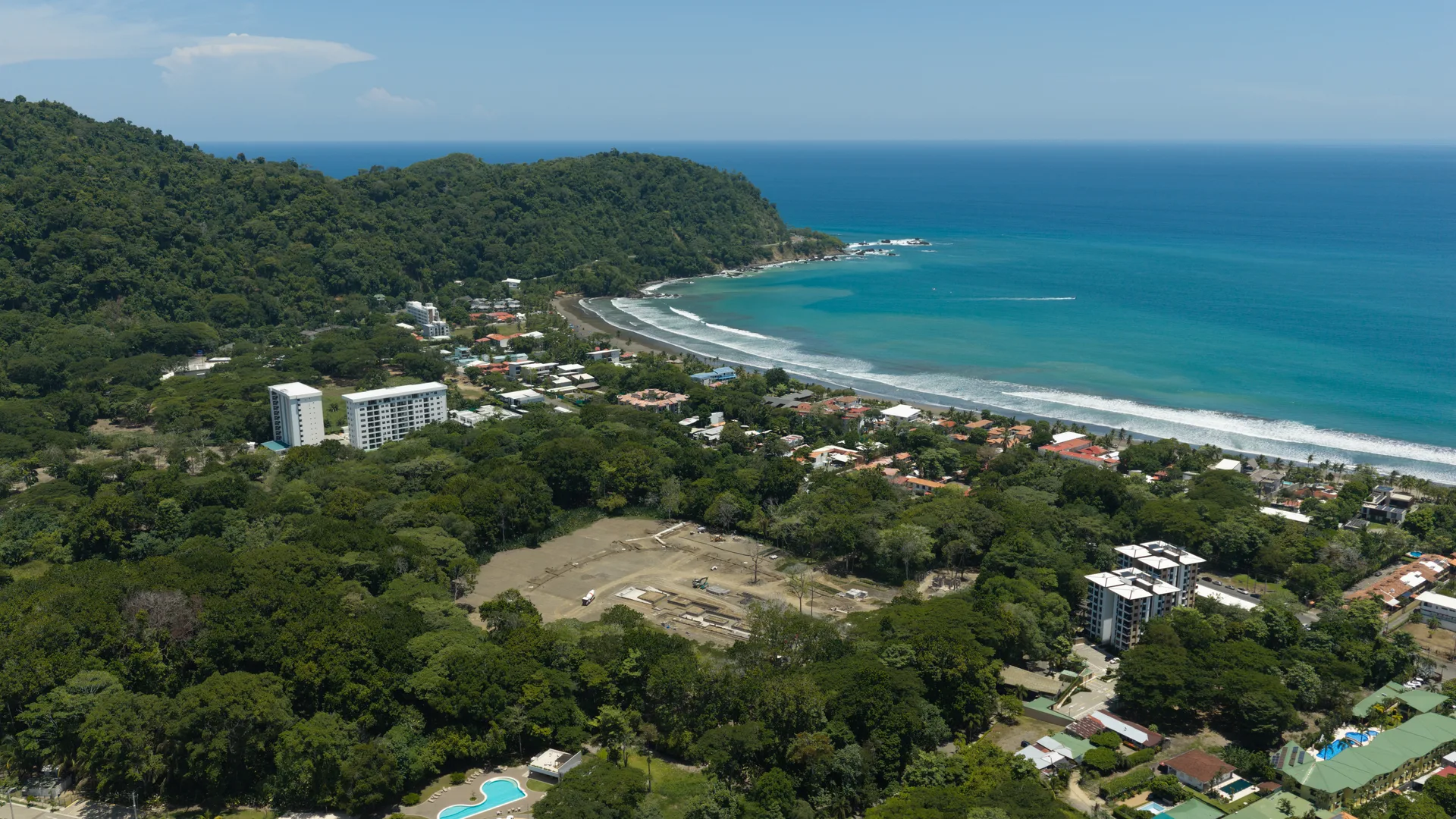 Drone photograph of the visible construction clearing amid forest, with Jacó Beach and the Pacific beyond.